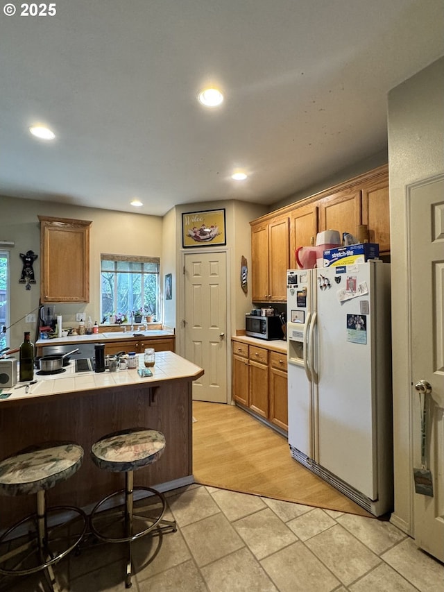 kitchen featuring white fridge with ice dispenser, tile counters, a breakfast bar area, stainless steel microwave, and brown cabinetry