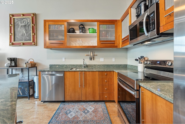 kitchen featuring dark stone countertops, brown cabinetry, a sink, glass insert cabinets, and appliances with stainless steel finishes