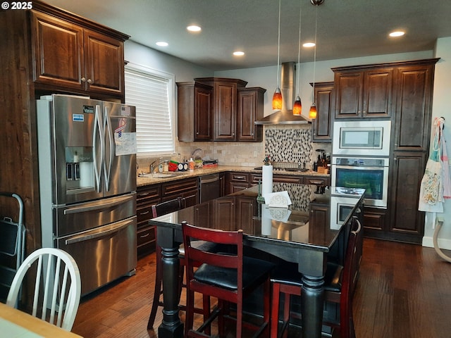 kitchen with pendant lighting, wall chimney range hood, stainless steel appliances, dark hardwood / wood-style floors, and a kitchen island