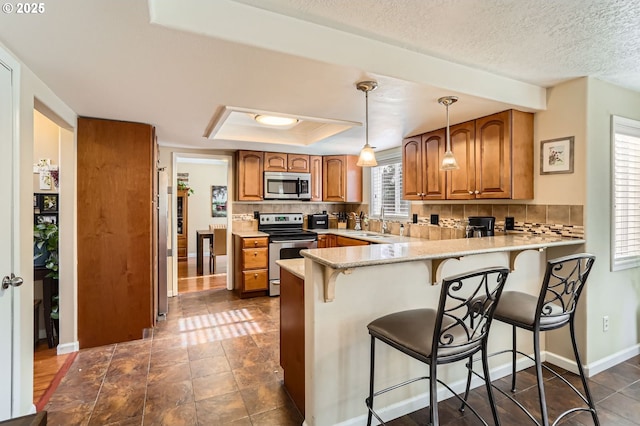 kitchen featuring brown cabinetry, decorative backsplash, a peninsula, a tray ceiling, and stainless steel appliances