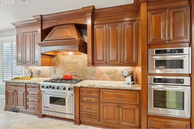 kitchen with light stone countertops, decorative backsplash, stainless steel appliances, brown cabinets, and premium range hood