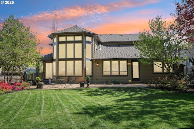 back of house at dusk featuring a lawn, fence, a shingled roof, and a patio area