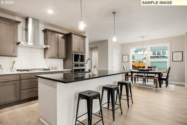 kitchen with appliances with stainless steel finishes, decorative backsplash, an island with sink, decorative light fixtures, and wall chimney exhaust hood