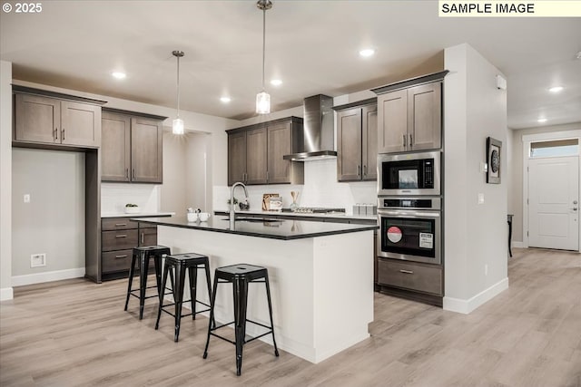 kitchen with pendant lighting, an island with sink, backsplash, stainless steel appliances, and wall chimney exhaust hood