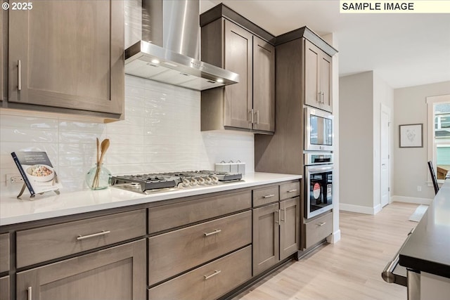 kitchen featuring wall chimney range hood, appliances with stainless steel finishes, dark brown cabinetry, decorative backsplash, and light wood-type flooring