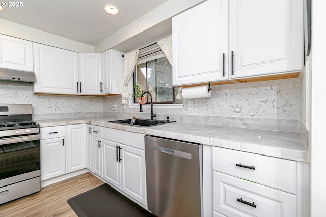 kitchen featuring light wood finished floors, a sink, stainless steel appliances, under cabinet range hood, and white cabinetry