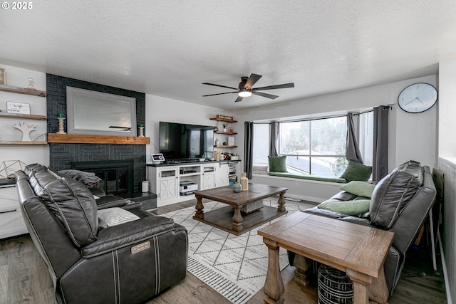 living area featuring a ceiling fan, a textured ceiling, wood finished floors, baseboards, and a brick fireplace