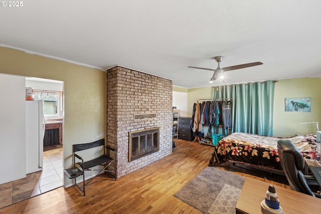 bedroom featuring wood finished floors, a fireplace, freestanding refrigerator, ceiling fan, and connected bathroom