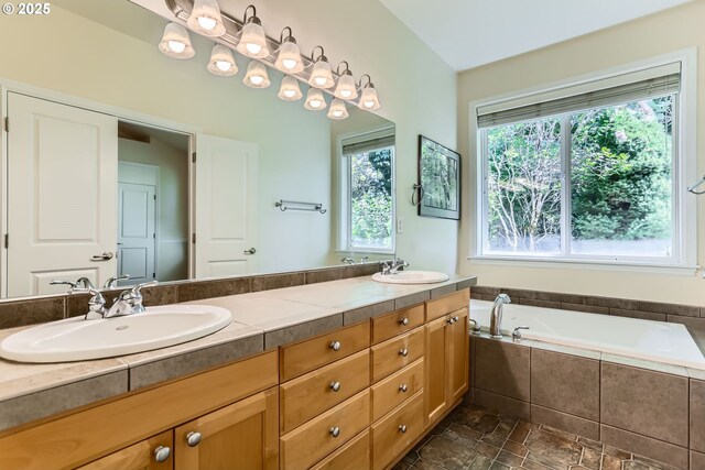 full bath featuring double vanity, tiled bath, and stone finish flooring