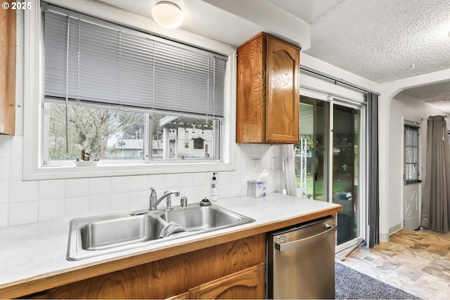 kitchen with backsplash, stainless steel dishwasher, brown cabinetry, a textured ceiling, and a sink