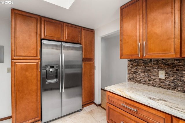 kitchen with light tile patterned flooring, light stone counters, brown cabinetry, backsplash, and stainless steel refrigerator with ice dispenser