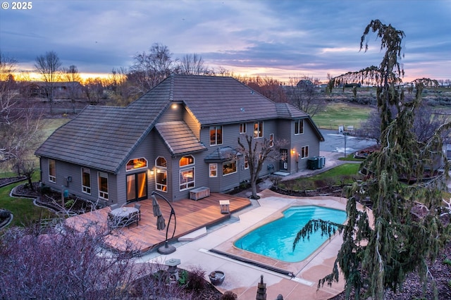 back of house at dusk with central AC unit, a tiled roof, a deck, and an outdoor pool