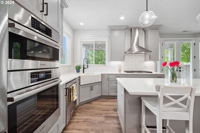 kitchen with gray cabinets, appliances with stainless steel finishes, wall chimney exhaust hood, a sink, and light wood-style flooring
