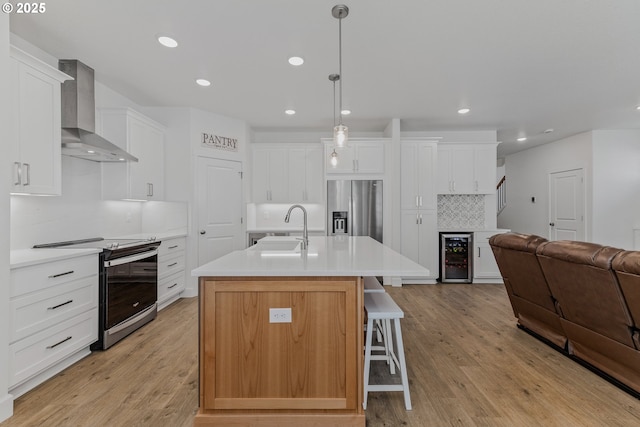 kitchen featuring stainless steel appliances, a sink, light countertops, wall chimney exhaust hood, and a center island with sink