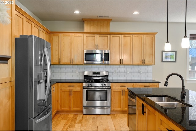 kitchen with backsplash, appliances with stainless steel finishes, dark stone counters, a sink, and light wood-type flooring