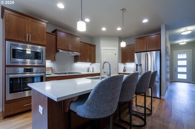 kitchen with a center island with sink, under cabinet range hood, a sink, backsplash, and appliances with stainless steel finishes