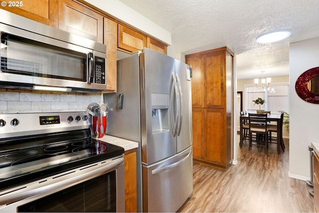 kitchen with stainless steel appliances, brown cabinetry, a chandelier, light wood-style flooring, and a textured ceiling