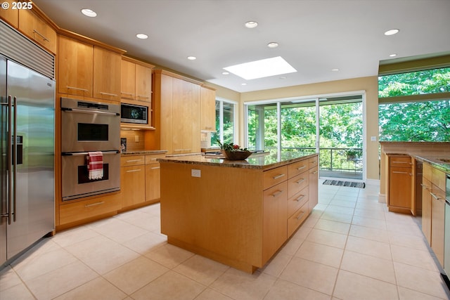 kitchen featuring built in appliances, a center island, a skylight, dark stone counters, and light tile patterned floors