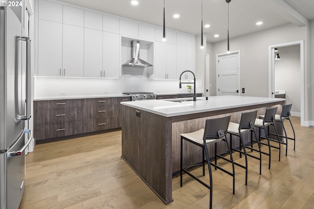 kitchen with high quality appliances, white cabinets, light countertops, wall chimney exhaust hood, and an island with sink