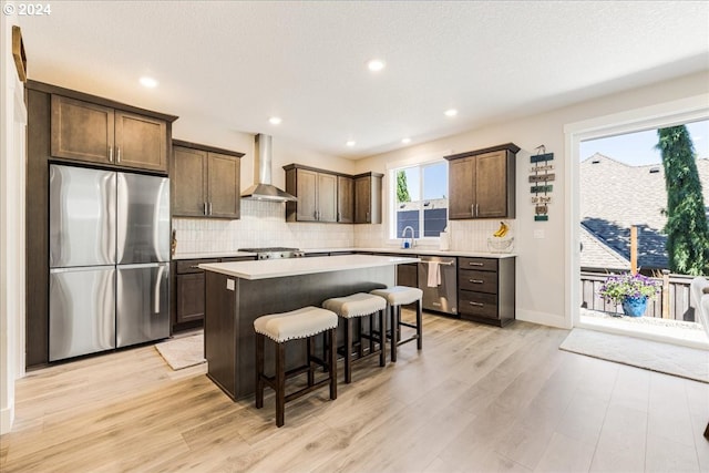 kitchen with light wood-type flooring, stainless steel appliances, a kitchen island, and wall chimney exhaust hood