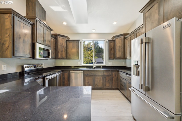 kitchen with stainless steel appliances, a sink, dark stone counters, dark brown cabinetry, and recessed lighting