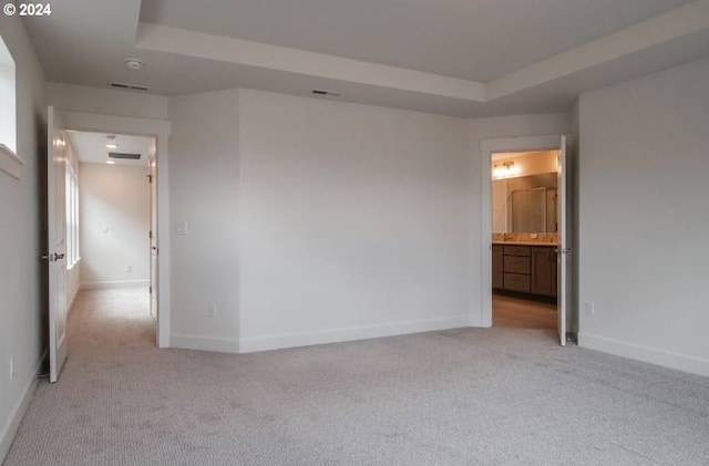 unfurnished bedroom featuring ensuite bath, light carpet, a tray ceiling, and baseboards