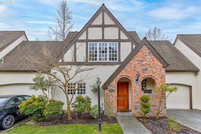 tudor home with an attached garage, brick siding, a shingled roof, driveway, and stucco siding