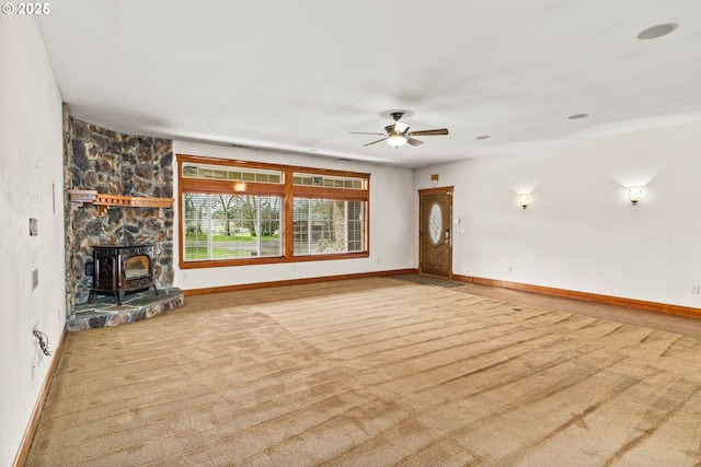 unfurnished living room featuring a wood stove, carpet, baseboards, and ceiling fan