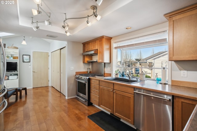 kitchen featuring light wood finished floors, brown cabinets, a sink, under cabinet range hood, and appliances with stainless steel finishes