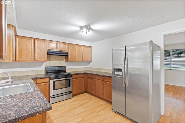 kitchen with a sink, under cabinet range hood, appliances with stainless steel finishes, dark countertops, and light wood-type flooring