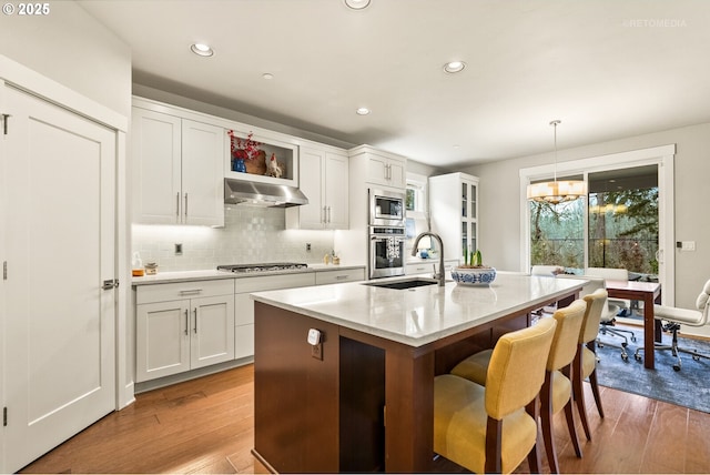 kitchen featuring a breakfast bar, a center island with sink, under cabinet range hood, wood finished floors, and stainless steel appliances