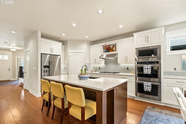 kitchen featuring backsplash, under cabinet range hood, light wood-style flooring, appliances with stainless steel finishes, and white cabinets