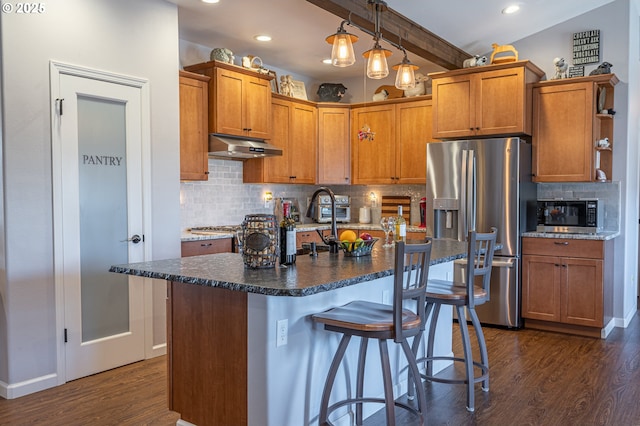 kitchen with dark wood-style flooring, beam ceiling, stainless steel appliances, under cabinet range hood, and a kitchen bar