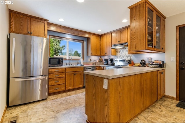 kitchen featuring under cabinet range hood, a peninsula, brown cabinetry, stainless steel appliances, and a sink