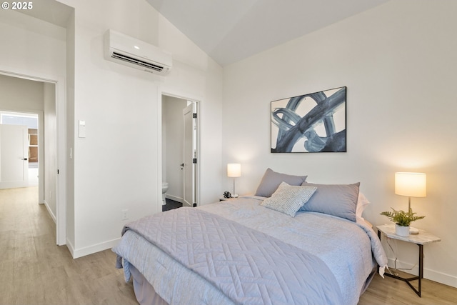 bedroom with ensuite bath, a wall mounted AC, vaulted ceiling, and light hardwood / wood-style flooring