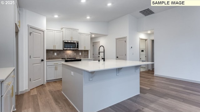 kitchen featuring a center island with sink, light countertops, stainless steel microwave, white cabinets, and vaulted ceiling