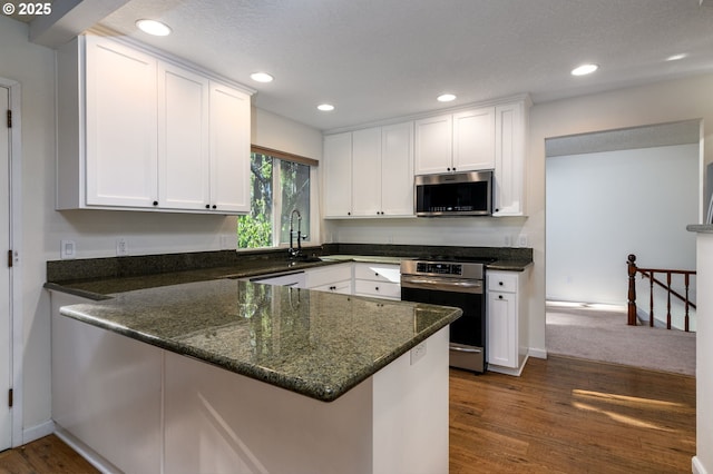 kitchen with dark wood-type flooring, appliances with stainless steel finishes, white cabinetry, dark stone countertops, and kitchen peninsula