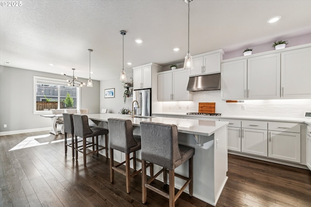 kitchen with a center island with sink, dark wood finished floors, decorative backsplash, stainless steel fridge with ice dispenser, and under cabinet range hood