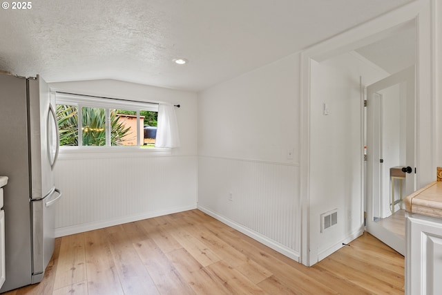 unfurnished room featuring vaulted ceiling, light wood-type flooring, a textured ceiling, baseboards, and a wainscoted wall