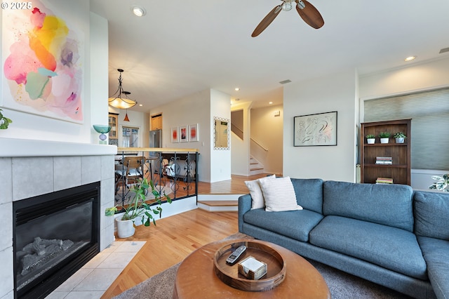 living room with light wood finished floors, a tiled fireplace, ceiling fan, stairway, and recessed lighting