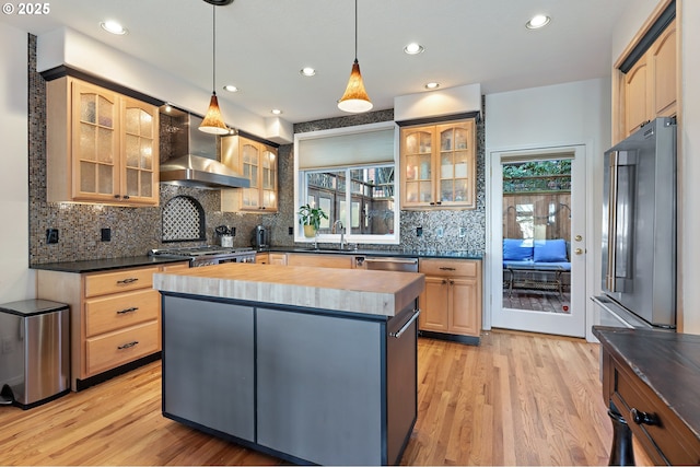 kitchen featuring decorative light fixtures, stainless steel appliances, glass insert cabinets, a kitchen island, and wall chimney range hood