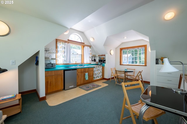 kitchen featuring stainless steel dishwasher, brown cabinetry, lofted ceiling, light colored carpet, and baseboards