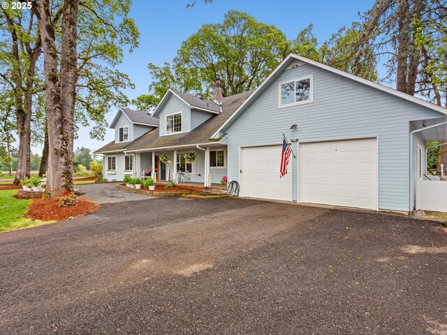 new england style home with covered porch, a chimney, aphalt driveway, an attached garage, and a shingled roof