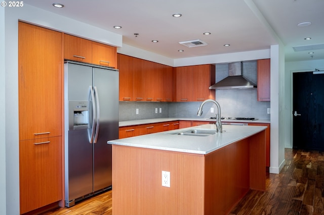 kitchen featuring sink, stainless steel fridge with ice dispenser, wall chimney exhaust hood, and an island with sink