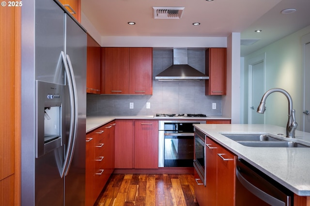 kitchen with wall chimney exhaust hood, dark wood-type flooring, stainless steel appliances, tasteful backsplash, and sink