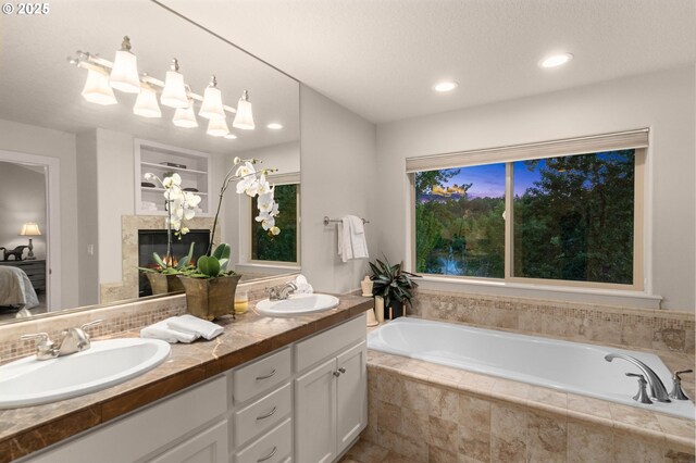 ensuite bathroom featuring a bath, double vanity, decorative backsplash, a glass covered fireplace, and recessed lighting