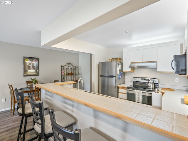 kitchen with tile counters, appliances with stainless steel finishes, a breakfast bar, under cabinet range hood, and white cabinetry