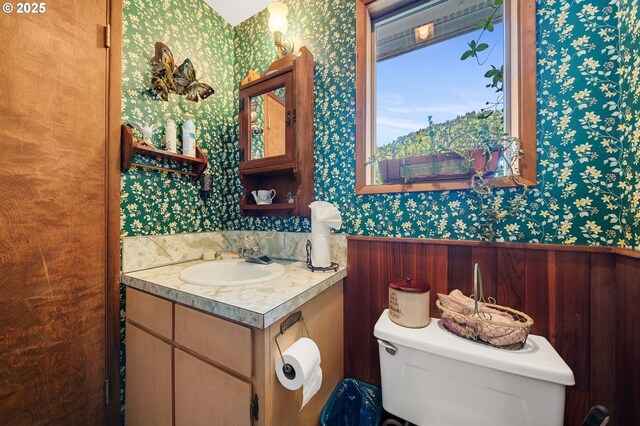bathroom with wainscoting, vanity, toilet, and wallpapered walls