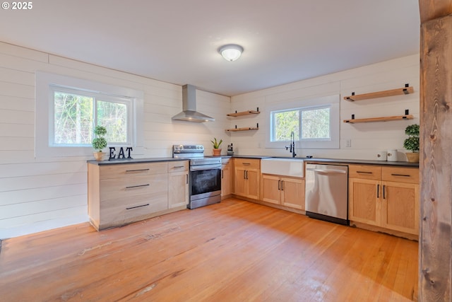 kitchen featuring stainless steel appliances, wall chimney range hood, light brown cabinets, light wood-type flooring, and sink