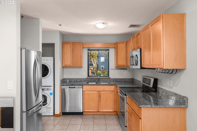 kitchen featuring sink, light tile patterned floors, stainless steel appliances, stacked washer / dryer, and dark stone counters